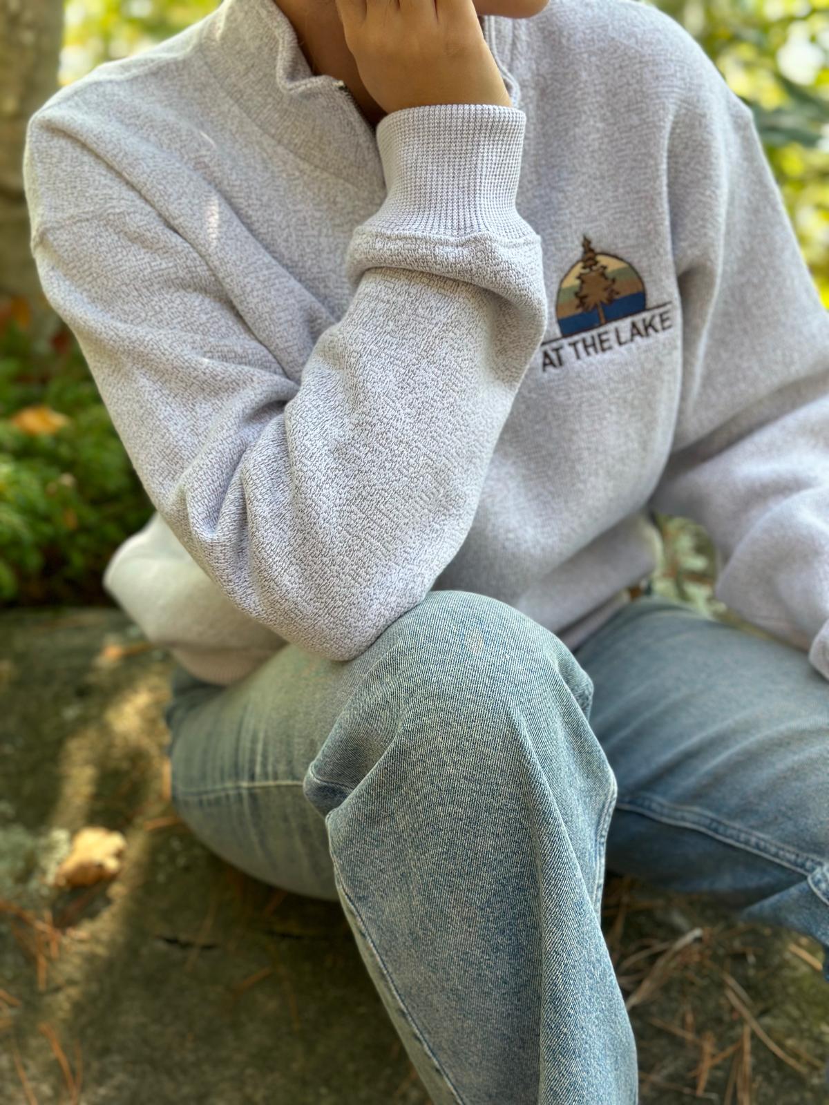 A person wearing a light grey quarter zip sweater with a logo on the chest sitting outdoors.