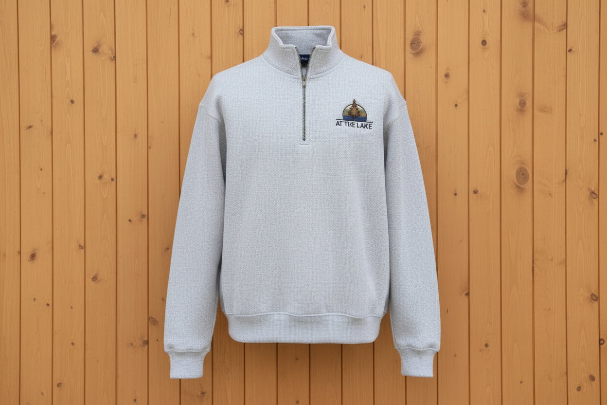 A person wearing a light grey quarter zip sweater with a logo on the chest sitting outdoors.